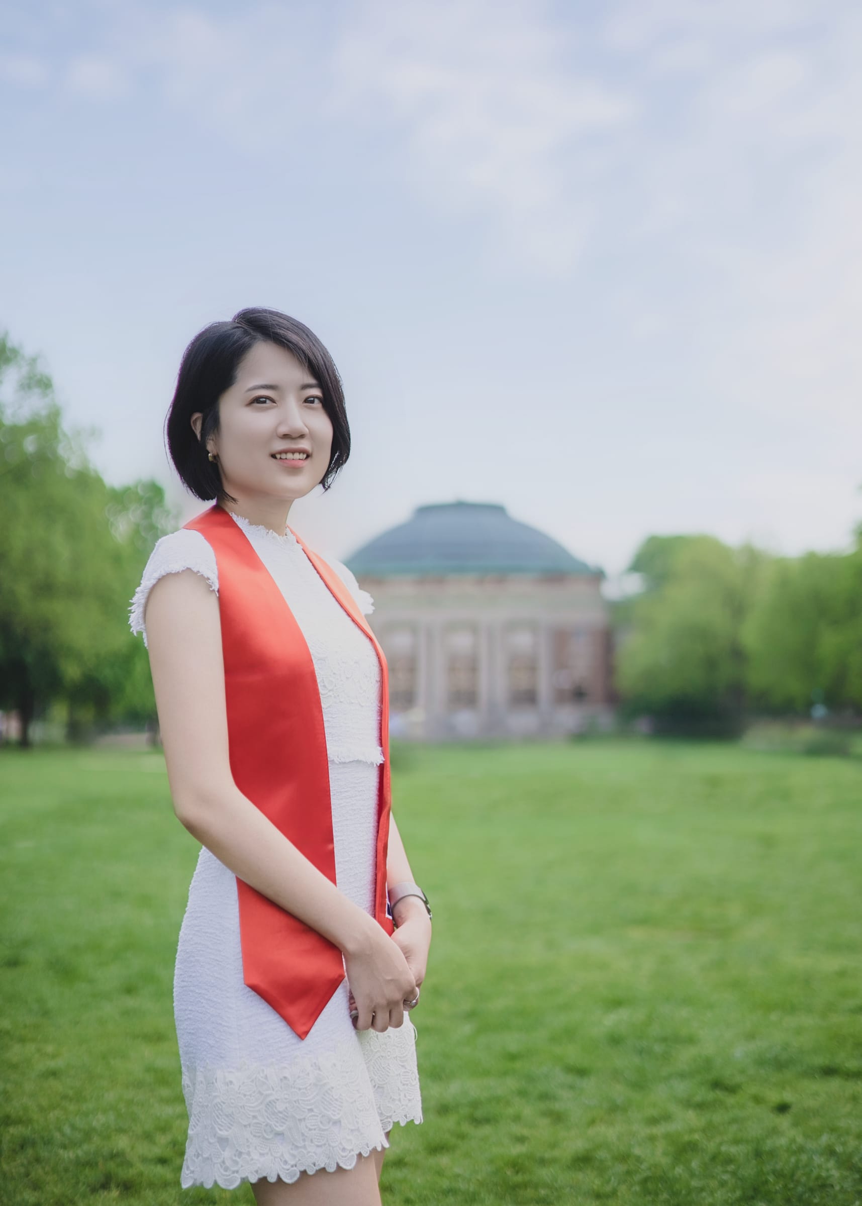Graduate in a white dress and red stole on a campus lawn