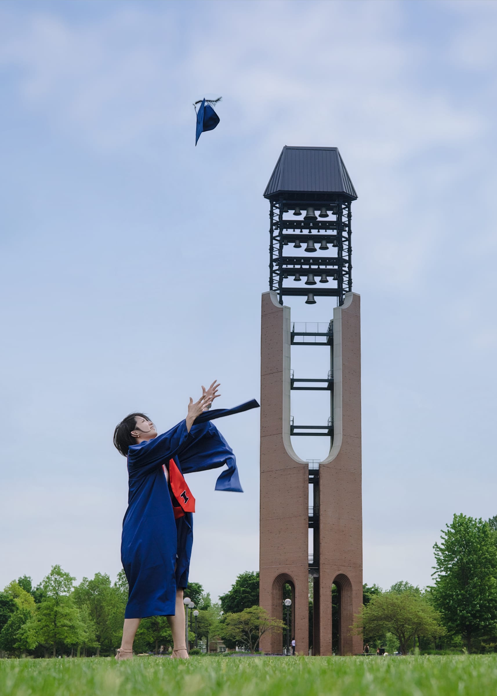 Graduate in a blue gown tossing a cap near a bell tower