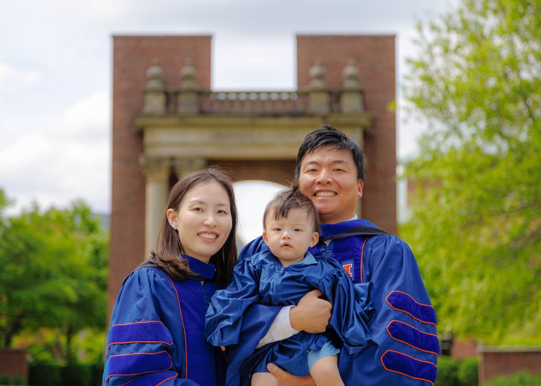 Graduating parents holding their child beneath a campus arch
