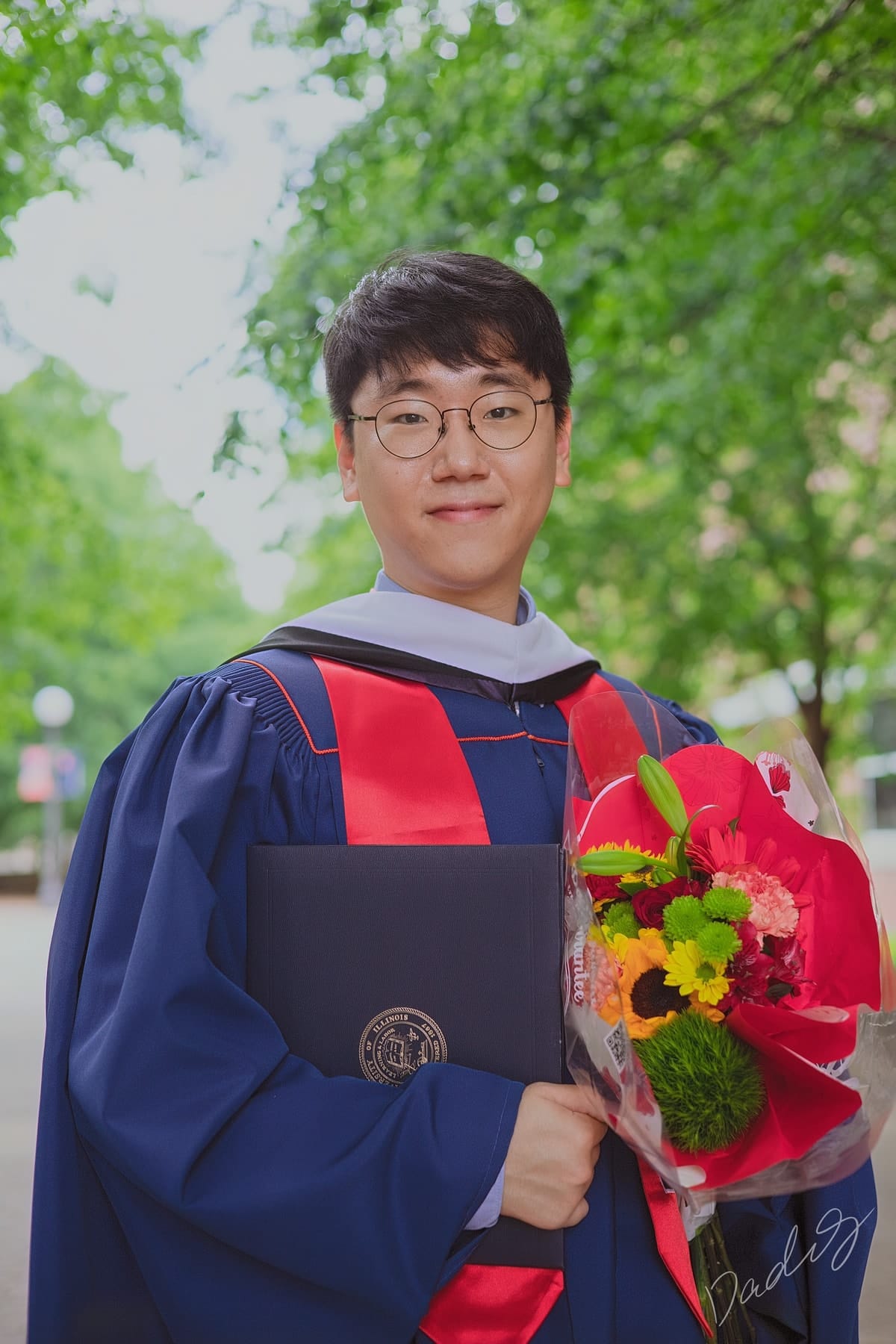 Graduate holding his diploma and a bouquet beneath green trees