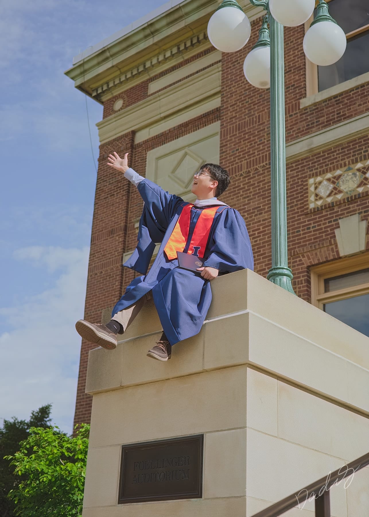 Graduate waving from a stone pedestal beside a brick building