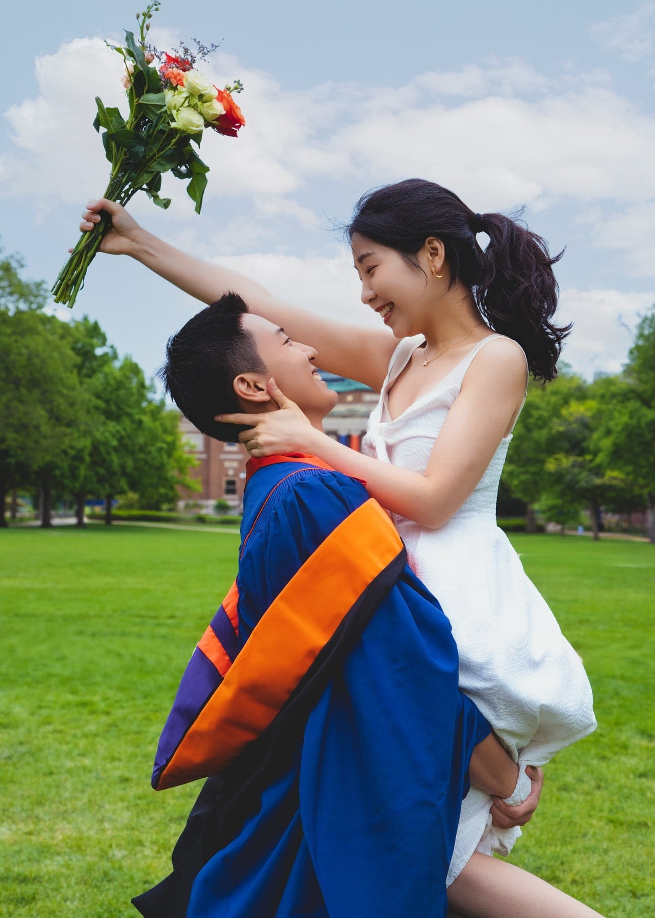 A graduate lifting his partner while she raises a bouquet